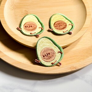 May include: Three embroidered avocado patches with smiling faces and tiny arms and legs. The patches are green, yellow, and brown, and are arranged on a wooden plate.