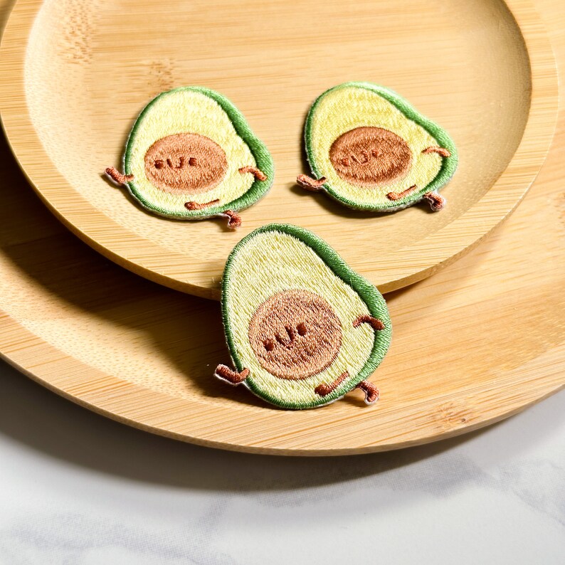 May include: Three embroidered avocado patches with smiling faces and tiny arms and legs. The patches are green, yellow, and brown, and are arranged on a wooden plate.