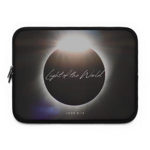 Puede incluir: Una funda rectangular negra para portátil con un gráfico de un eclipse solar con un halo de luz blanca brillante. El texto Light of the World está escrito en una fuente cursiva blanca en el centro, con el texto JOHN 8:12 impreso debajo en blanco.