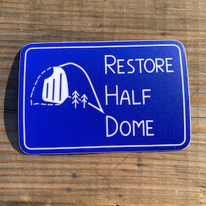 May include: A rectangular blue sticker with a white border featuring a white line drawing of a mountain dome and two small pine trees. The text RESTORE HALF DOME is printed in white capital letters on the right side of the sticker design.