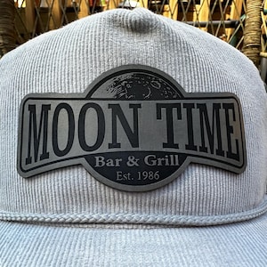 May include: A light gray corduroy hat with a black patch. The patch features the words "MOON TIME" above "Bar & Grill" and "Est. 1986". A detailed illustration of the moon is above the words "Bar & Grill".