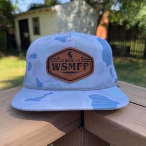 May include: A camouflage print cap in white and blue, with a flat peak. It has a brown leather patch displaying the text "ATHENS, GA" and "WSMFP". The cap is finished with a grey rope detail around the crown.