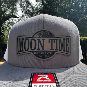 May include: Grey flat-peak cap with white mesh at the back. The cap has a rectangular patch with the words "MOON TIME Bar & Grill Est. 1986". A red and white tag with the brand name "RICHARDSON" is visible.