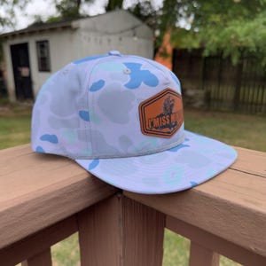 May include: A flat-brimmed baseball cap with a white and blue camouflage pattern. It has a brown leather patch with the text "I Miss Mike" and a grey rope detail. The cap is displayed on a wooden surface.