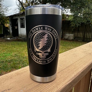 May include: Black stainless steel tumbler with a silver rim and lid. The tumbler features an engraved design with a skull and lightning bolt graphic, and the text "THE BOTTLE WAS DUSTY BUT THE LIQUOR WAS CLEAN".