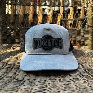 Peut inclure: Casquette de camionneur grise en velours côtelé avec un dos en filet noir. Le chapeau présente un écusson rectangulaire noir avec les mots "MOON TIME Bar & Grill Est. 1986". La casquette a une visière incurvée.