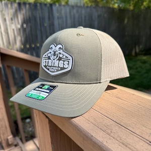 Può includere: Cappellino da camionista verde oliva con retro in rete e visiera curva. Il cappello presenta una toppa con la scritta "STRINGS" e una grafica di capra. C'è un'etichetta Richardson 112RE.