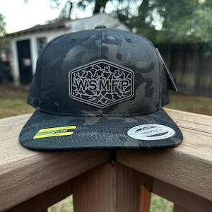 May include: A black and green camouflage snapback hat with a flat brim. The hat features a patch with the letters "WSMFP" in a silver color. The hat has a "MultiCam" sticker on the brim.
