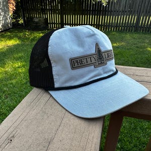 Pode incluir: Boné de caminhoneiro azul claro com parte traseira em malha preta e aba curva. O boné apresenta um patch retangular com as palavras "PRETTY LIGHT" e um gráfico de árvore. Um detalhe de corda preta acentua a aba.