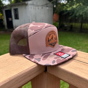 Puede incluir: Una gorra de camionero con camuflaje rosa y marrón, parte trasera de malla marrón y visera plana. Un parche de cuero en el frente muestra un gráfico de montaña y el texto Climb To Safety Mountain Rescue Squad. Tiene una pegatina en la visera.