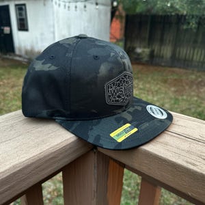 Peut inclure: Casquette de baseball camouflage noir et vert avec une visière plate. Le chapeau comporte un écusson avec les lettres "WSM" et un petit autocollant indiquant "MultiCam".