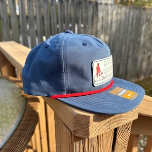 Puede incluir: Gorra de béisbol azul marino con visera plana y detalle de cuerda roja. La gorra presenta un parche rectangular con el texto "GO TO CHURCH Or the Devil wins" y un gráfico de una persona. Fabricada en tejido resistente.