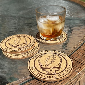 May include: Four brown cork coasters with a Grateful Dead skull and lightning bolt design. The coasters have the text "The bottle was dusty but the liquor was clean" around the design. The coasters are on a glass table with a glass of ice water.