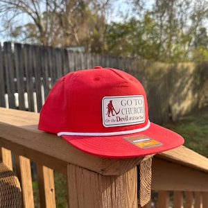 Puede incluir: Gorra de béisbol roja con detalle de cuerda blanca y visera plana. La gorra presenta un parche rectangular con el texto "GO TO CHURCH Or the Devil Will Get You" y un gráfico de una persona con un palo de hockey.
