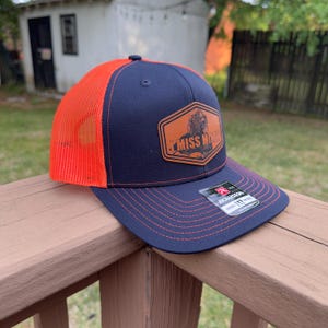 May include: A navy blue and orange trucker hat with a patch that reads "MISS MIKE". The hat has a curved brim and a mesh back. The patch is a brown colour and has a picture of a person on it. The hat is sitting on a wooden surface.