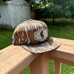 Puede incluir: Una gorra de béisbol con un patrón de camuflaje marrón y beige, una corona estructurada y una visera plana. Un detalle de cuerda negra se sitúa sobre la visera. Un parche hexagonal blanco en la parte frontal muestra la silueta negra de una persona sentada.