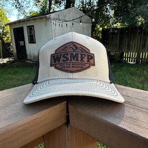 May include: A beige and black trucker hat with a brown leather patch. The patch features the text "WSMFP" above "KISS THE MOUNTAIN AIR WE BREATHE" and a mountain graphic. The hat is sitting on a wooden railing.