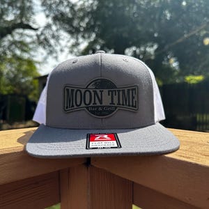 Peut inclure: Casquette grise et blanche avec l'inscription "MOON TIME Bar & Grill Est. 1998" sur le devant. La casquette a une visière plate et une fermeture à pression. Le chapeau est posé sur une balustrade en bois.
