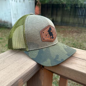 Peut inclure: Casquette de camionneur avec une visière camouflage et une couronne beige. Le chapeau présente un écusson en cuir marron avec une silhouette d'une personne pêchant et plusieurs oiseaux. Le dos en filet est vert olive.