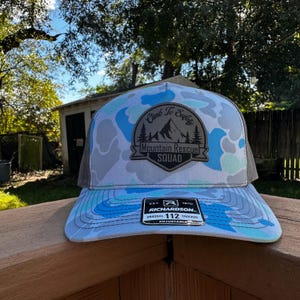 Peut inclure: Casquette de camionneur avec un motif camouflage dans les tons de bleu, gris et blanc. Le chapeau comporte un écusson avec le texte "Climb To Safety Mountain Rescue Squad". La visière est incurvée et l'arrière est en mesh.