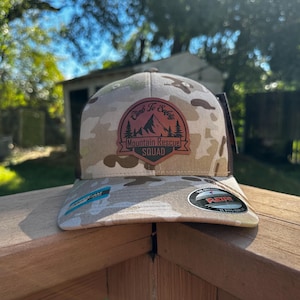 Peut inclure: Casquette de baseball camouflage avec un dos en filet noir. La casquette présente un écusson avec le texte "Climb to Safety Mountain Rescue Squad" et un graphique de montagne. La visière a un autocollant Flexfit.