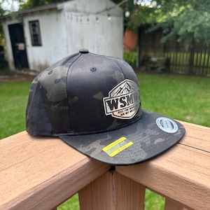 Op de afbeelding: Een zwarte multicam baseballpet met een zilverkleurige zeshoekige patch op de voorkant met een bergafbeelding en de tekst WSMR KISS THE MOUNTAIN AIR WE BREATHE. De platte klep is voorzien van een gele en zwarte Multicam Black sticker.
