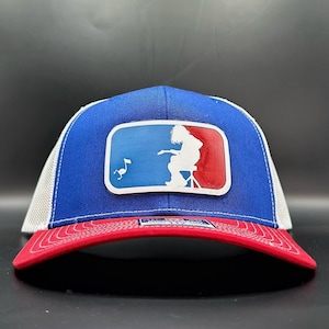Peut inclure: Casquette de baseball bleue et rouge avec un dos en maille blanche. L'avant présente un écusson rectangulaire avec une silhouette de musicien et une note de musique. La visière est rouge et la couronne est bleue.