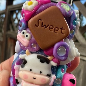 Könnte beinhalten: Ein individuell dekorierter Feuerzeug mit Kuh-, Blumen- und Alien-Design. Das Feuerzeug hat ein braunes Quadrat mit dem Wort "Sweet" darauf. Das Feuerzeug ist mit rosa, lila, weißen und blauen Farben verziert.