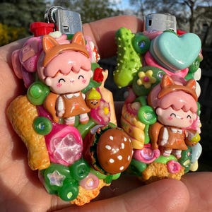 Puede incluir: Dos encendedores decorativos con diseños esculpidos coloridos. Cada encendedor presenta un personaje de dibujos animados con cabello rosado y una diadema de orejas de zorro, rodeado de adornos temáticos de comida.