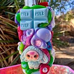May include: A Bic lighter decorated with colorful clay embellishments, including a light blue chocolate bar with the word MILK embossed on it, a small light blue sheep character, and various purple, green, and pink shapes with glittery accents.