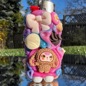 May include: A colorful, decorative lighter with a whimsical design. The lighter is covered in various shapes, including bows, flowers, and a cartoon character. The color palette includes pink, purple, and white, with a silver metal top.