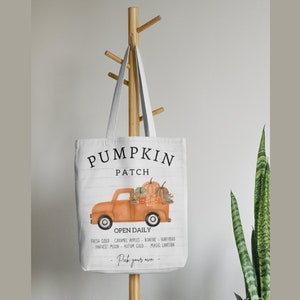 May include: White tote bag with a pumpkin patch design. The bag features an orange truck filled with pumpkins and the words "Pumpkin Patch" in black. Additional text includes "Open Daily" and a list of fall-themed items.