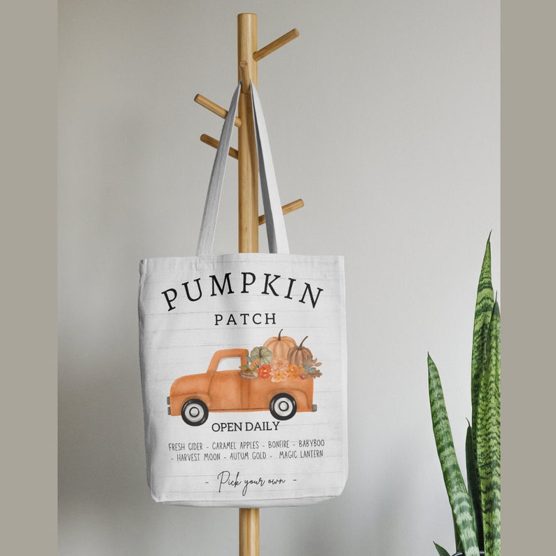 May include: White tote bag with a pumpkin patch design. The bag features an orange truck filled with pumpkins and the words "Pumpkin Patch" in black. Additional text includes "Open Daily" and a list of fall-themed items.