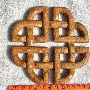 May include: A carved wooden Celtic knot in a natural wood tone. The knot is a four-part design with rounded edges and visible wood grain. The piece is approximately 5 inches wide.