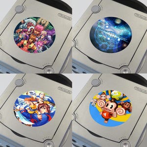 May include: Four silver Nintendo GameCube consoles with different circular game art stickers. The stickers feature colorful illustrations of characters and game titles, including "Fantasy Star Online" and a monkey character.