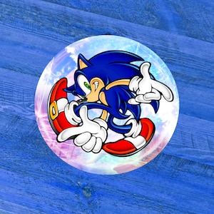 May include: A round button featuring the animated character Sonic the Hedgehog. The character is blue with white gloves and red shoes, set against a colorful, swirling background. The button is on a blue wooden surface.