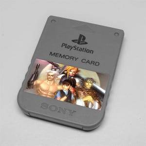 May include: A gray PlayStation memory card with the PlayStation logo and the words "MEMORY CARD" printed on it. The card features a colorful image of four characters. The word "SONY" is printed at the bottom.