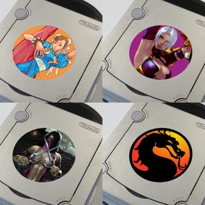 Könnte beinhalten: Vier verschiedene Videospiel-Charakterdesigns, die auf runde Aufkleber gedruckt sind. Die Aufkleber befinden sich auf der Oberseite von vier silbernen Nintendo GameCube-Konsolen. Die Charaktere sind Chun-Li aus Street Fighter, eine weibliche Figur aus der Dead or Alive-Serie, eine weibliche Figur aus der Mortal Kombat-Serie und das Mortal Kombat-Drachenlogo.