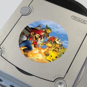 May include: A silver Nintendo GameCube console with a sticker featuring Mario, Luigi, Bowser, and Pikachu characters from the Super Smash Bros. video game.