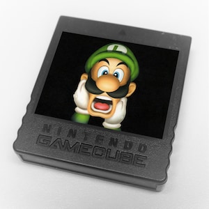 Puede incluir: Un cartucho negro de Nintendo GameCube con una imagen colorida de Luigi con expresión de sorpresa. El personaje lleva un gorro y guantes verdes, con un gran bigote y la boca abierta. El texto "NINTENDO GAMECUBE" está impreso en el cartucho.