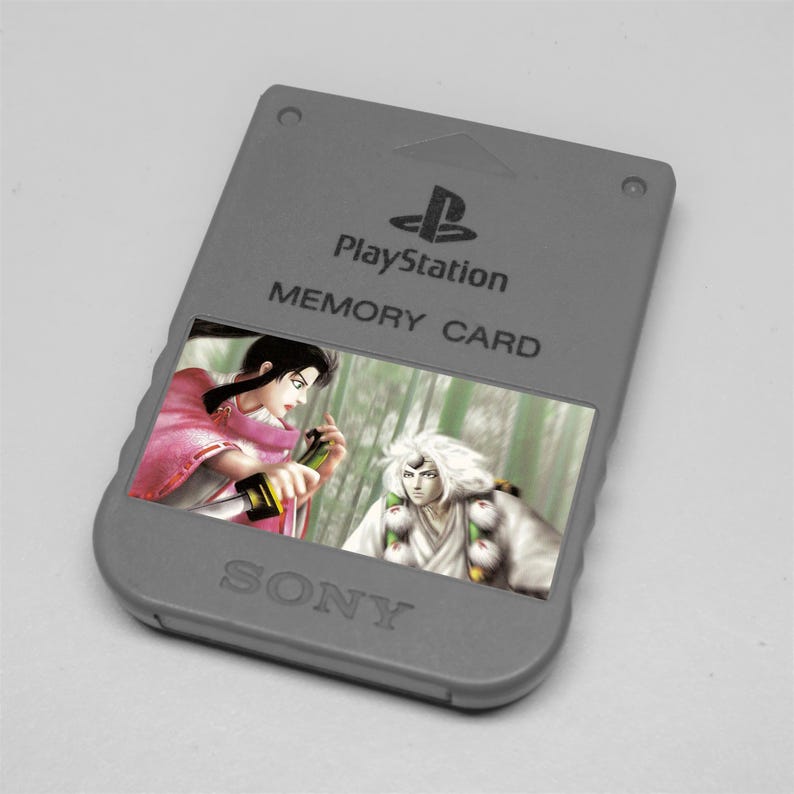 May include: A gray PlayStation memory card with the PlayStation logo and the word "MEMORY CARD" printed on it. The card features a colorful image of two anime characters. The word "SONY" is printed at the bottom.