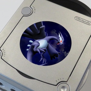 May include: A silver Nintendo GameCube console featuring a circular sticker on its top surface. The sticker displays a dark purple, winged creature with red eyes against a dark blue, cloudy sky background. The console has visible Power, Reset, and Open buttons.