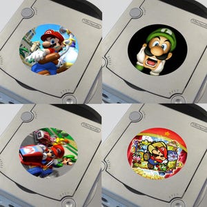 May include: Four silver GameCube consoles with different circular stickers featuring characters from the Mario franchise. The stickers feature Mario, Luigi, Wario, and a group of Mario characters.