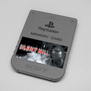 May include: A grey PlayStation memory card with the PlayStation logo and the text "MEMORY CARD". The card features a graphic from the video game "SILENT HILL" with the game's title in red.