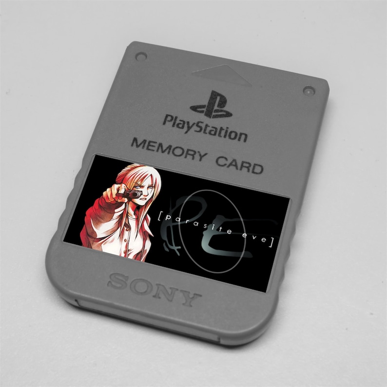 May include: Gray PlayStation memory card with a colorful graphic of a woman pointing a gun, with the text "[parasite eve]" on a black background. The card has the PlayStation logo and the words "PlayStation" and "Memory Card" printed on it. The word "SONY" is printed at the bottom.