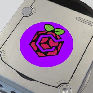 May include: A purple circle sticker with a black and red raspberry pi logo on a silver Nintendo GameCube console. The logo has a green leaf on top.