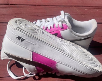 May include: White leather sneakers with a pink and black design. The sneakers have a black swoosh logo and the word "AIR" printed on the back.