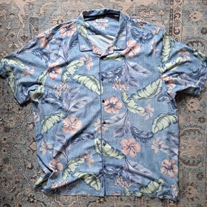 K&ouml;nnte beinhalten: Ein hellblaues Hemd mit tropischem Blumenmuster. Das Hemd hat kurze &Auml;rmel und einen Kragen. Das Design umfasst rosa Hibiskusbl&uuml;ten, gr&uuml;ne Bl&auml;tter und violettes Laub.