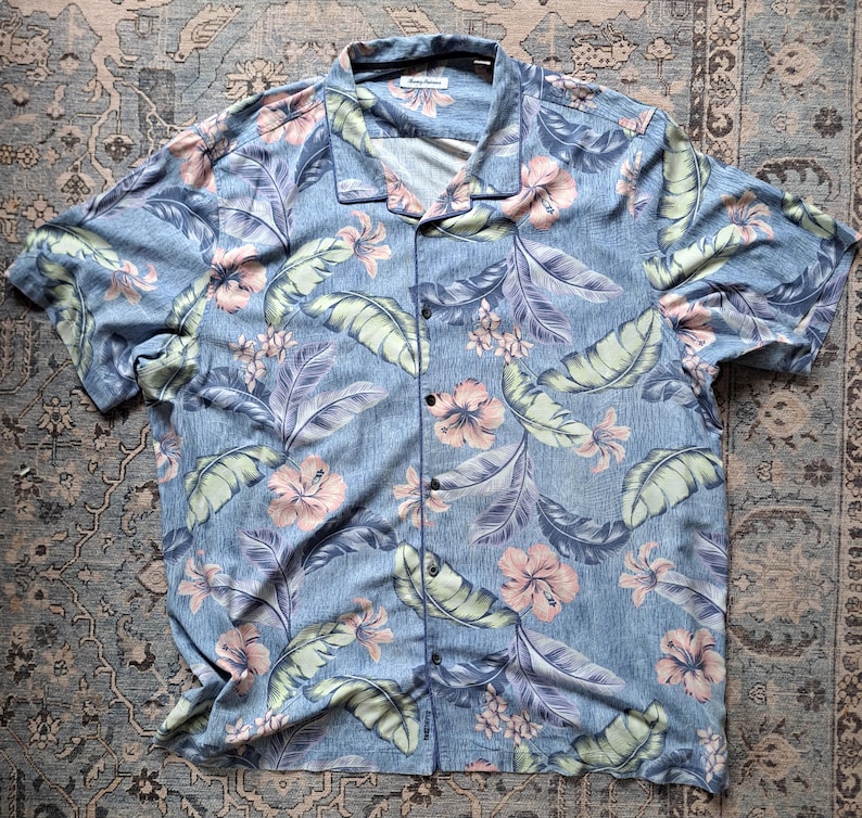 K&ouml;nnte beinhalten: Ein hellblaues Hemd mit tropischem Blumenmuster. Das Hemd hat kurze &Auml;rmel und einen Kragen. Das Design umfasst rosa Hibiskusbl&uuml;ten, gr&uuml;ne Bl&auml;tter und violettes Laub.