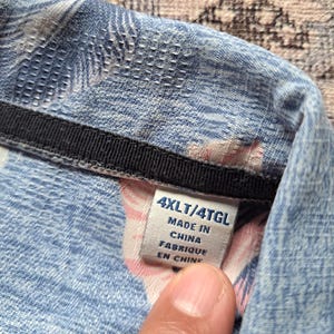 K&ouml;nnte beinhalten: Nahaufnahme eines blauen Hemdes mit floralem Muster. Das Etikett lautet "4XLT/4TGL" und "Made in China". Der Stoff hat eine strukturierte Optik mit einem schwarzen Kragendetail. Das Etikett sagt auch "Fabriqu&eacute; en Chine".